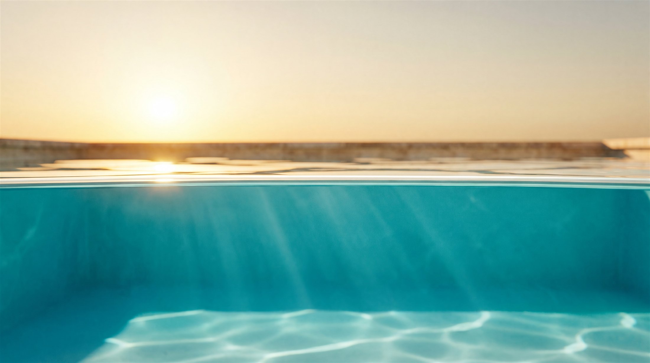The waterline of a glass-clear pool at golden hour, seen half above and half below the surface